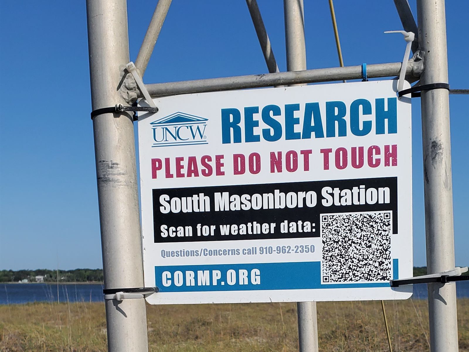 Sign on a weather tower reading UNCW Research, Please Do Not Touch, South Masonboro Station, Scan for weather data, cormp.org